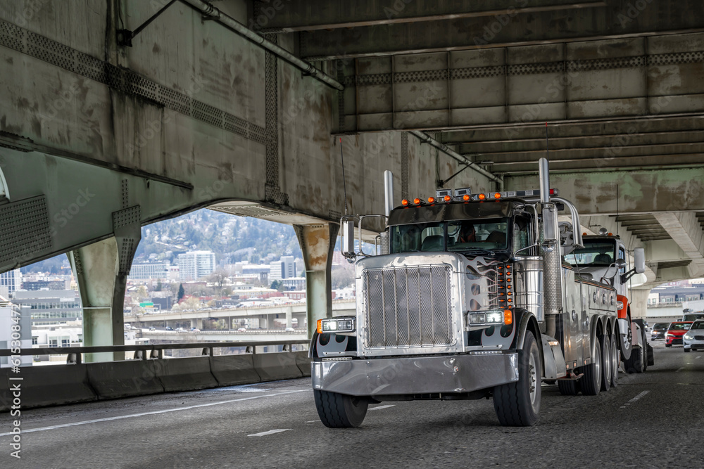 Big rig black towing semi truck tow another broken semi truck without semi trailer running with turned on headlights on the transportation Fremont bridge in Portland downtown Big rig black towing semi truck tow another broken semi truck without semi trailer running with turned on headlights on the transportation Fremont bridge in Portland downtown