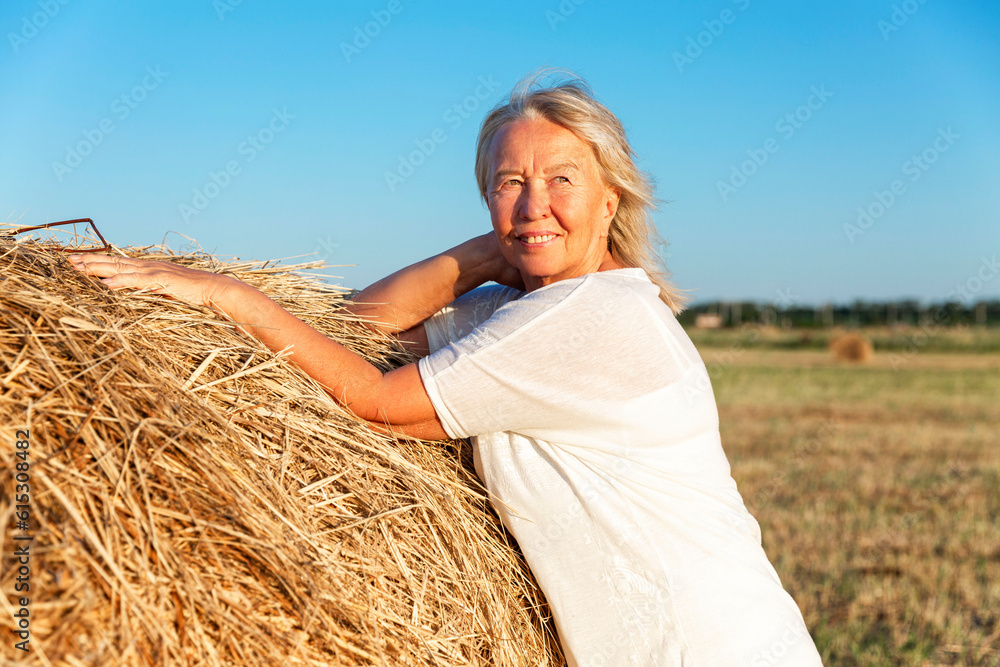 Smiling elderly woman in a white sweater and trousers in a field with hay rolls at sunset. Farming, freedom and nature. activity and longevity. Smiling elderly woman in a white sweater and trousers in a field with hay rolls at sunset. Farming, freedom and nature. activity and longevity.