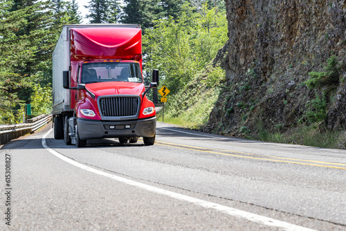 Bright red big rig semi truck with roof spoiler transporting cargo in dry van semi trailer driving on the winding mountain road with forest and rock wall on the sides