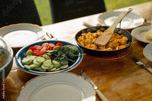 Green cut salads and meat omelet in background on picnic breakfast table outside, with white dishes