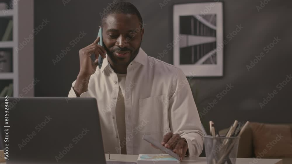 Waist up of young African American businessman talking on mobile phone while sitting at office desk against laptop computer Waist up of young African American businessman talking on mobile phone while sitting at office desk against laptop computer
