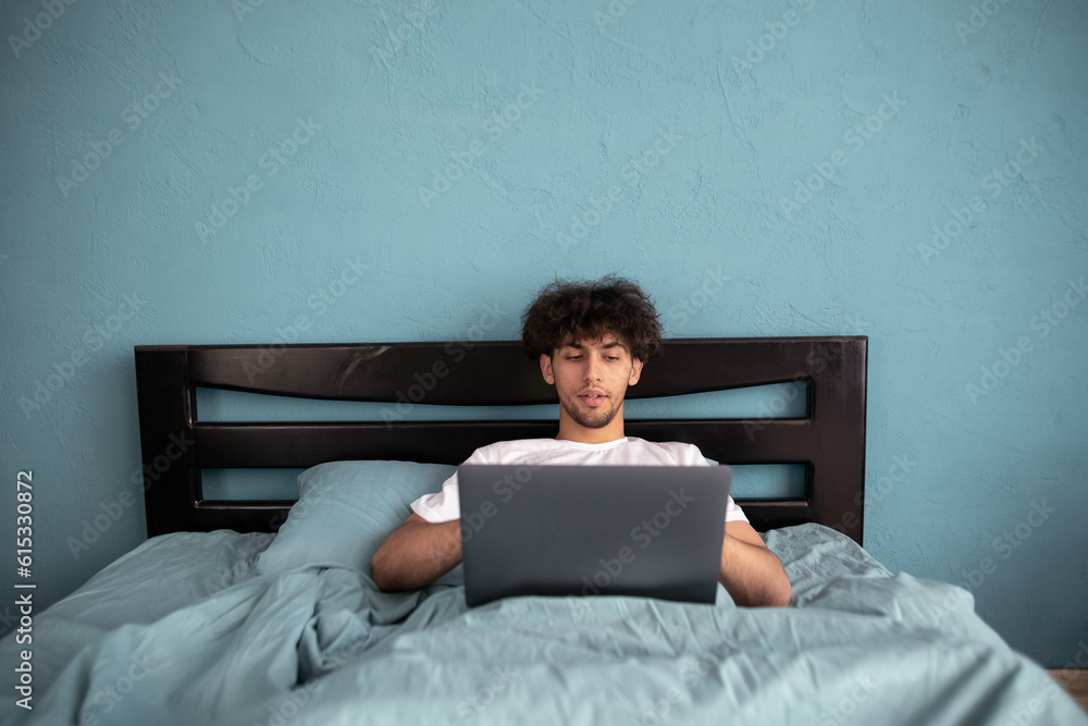 Young man working using laptop lying in bed in the morning, overtime remote work on weekend, freelancer working at home Young man working using laptop lying in bed in the morning, overtime remote work on weekend, freelancer working at home