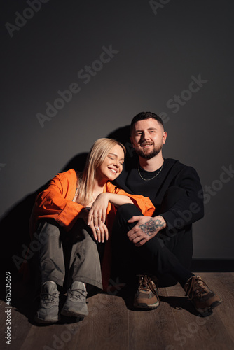 Full body of smiling blond haired woman in stylish clothes leaning on knee of boyfriend while sitting together against black wall. Vertical photo