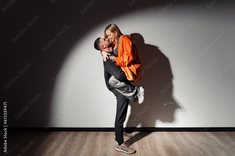 Full body side view of young man lifting up and embracing woman while touching noses in the light of a spotlight in dark room Full body side view of young man lifting up and embracing woman while touching noses in the light of a spotlight in dark room