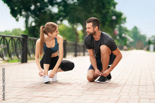 Image of young happy smiling couple, woman training with man or bearded coach trainer tying shoe laces, before morning jogging, cardio workout outdoors. Fitness, sport, healthy lifestyle concept.