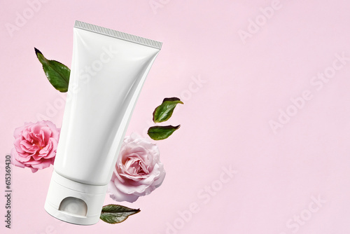 White empty cosmetic tube, on a pink background with delicate pink roses. Natural cosmetics with rose extract or skin care. Flat lay, top view, copy space.