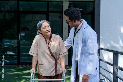 Asian elderly woman walking with walker under close supervision of physiotherapist in rehab center