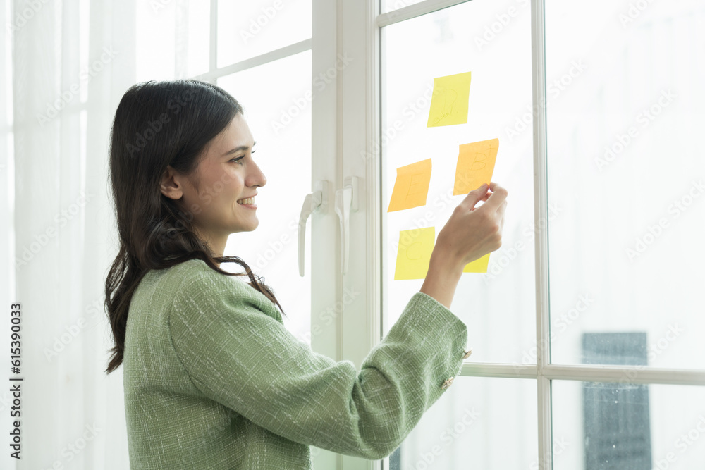 Happy young business woman working with post it note or sticky note on glass wall in office. Business people and office concept
