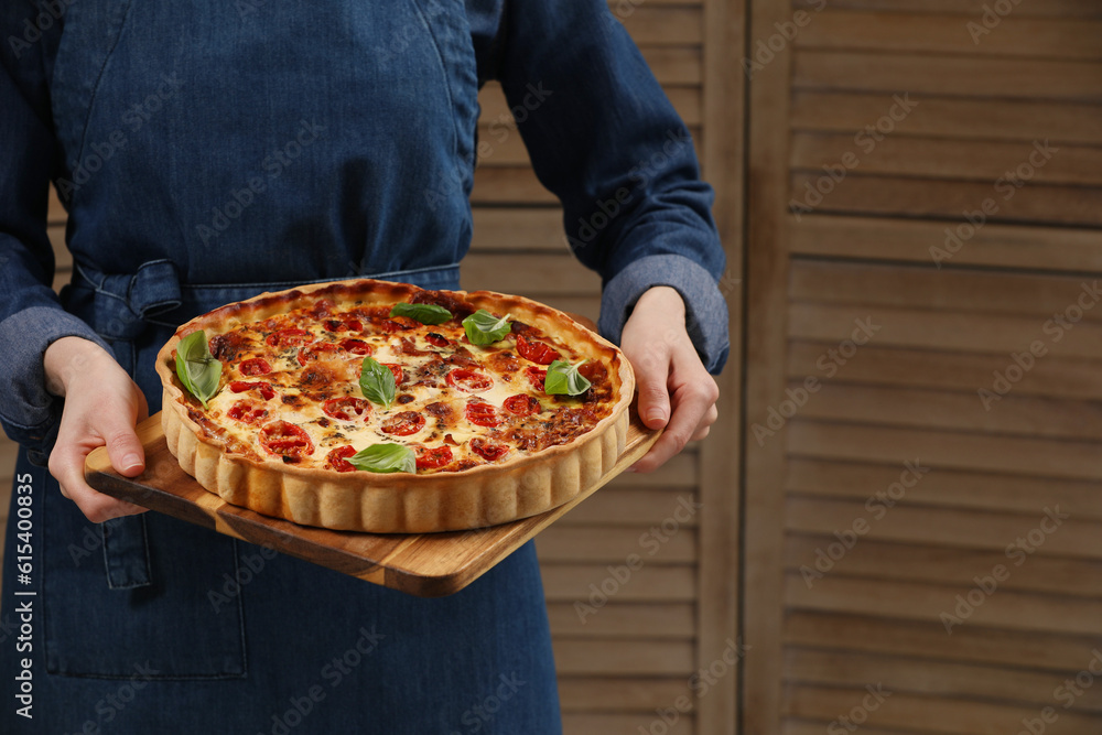 Woman holding delicious homemade quiche with prosciutto against wooden folding screen, closeup. Space for text