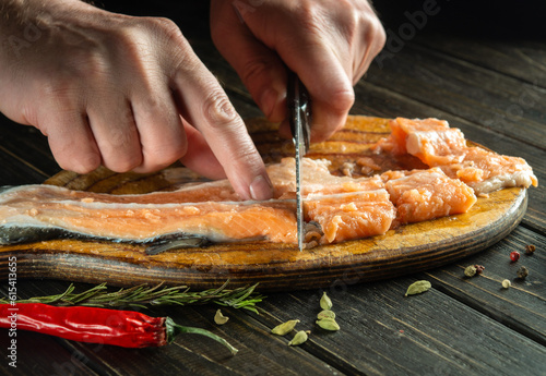 The chef cuts fresh salmon fish with a knife on a kitchen cutting board. Cooking a delicious fish dish according to an old recipe with the addition of spices and pepper