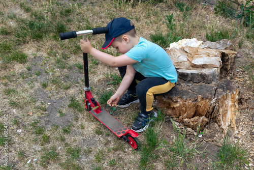 A little boy with a red scooter sits on an old stump