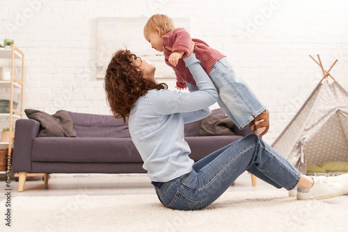 quality family time, curly woman lifting toddler daughter and sitting on carpet in cozy living room, work life balance, denim clothes, casual attire, family relationships, modern parenting