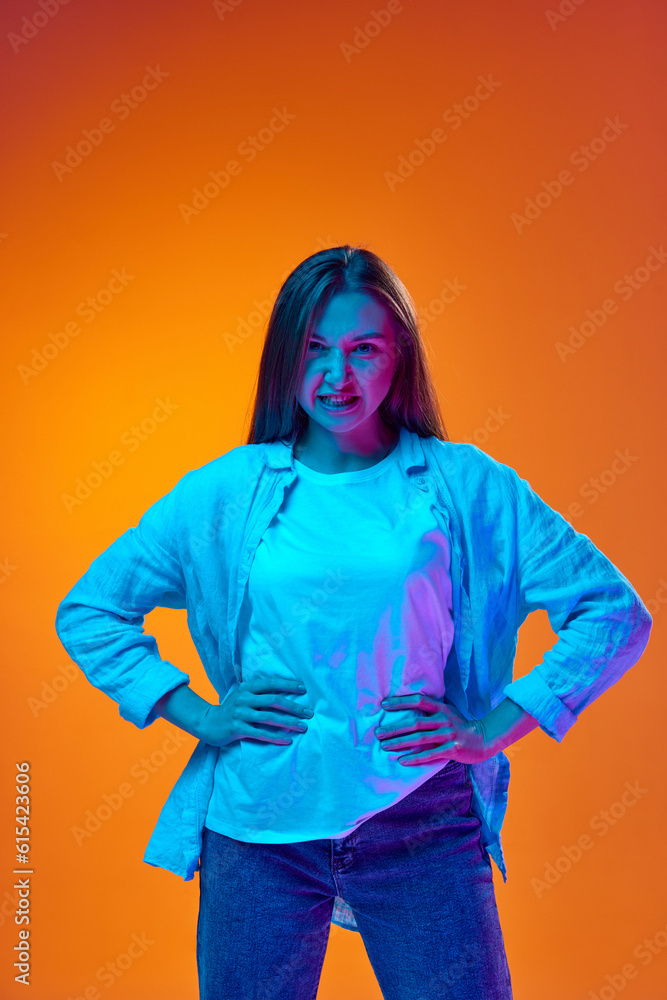 Portrait of young woman in casual clothes standing with angry face against gradient orange studio background in neon light. Irritation