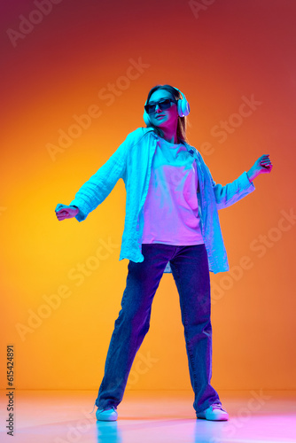 Young woman in casual clothes and sunglasses listening to music in headphones against gradient orange studio background in neon light © Lustre Art Group 