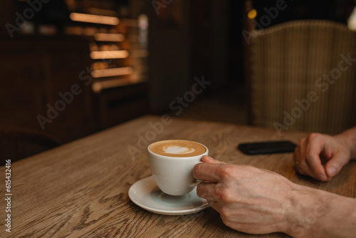 Close-up of man holding a cup of cappuccino on wooden table in cafe. Copy space.