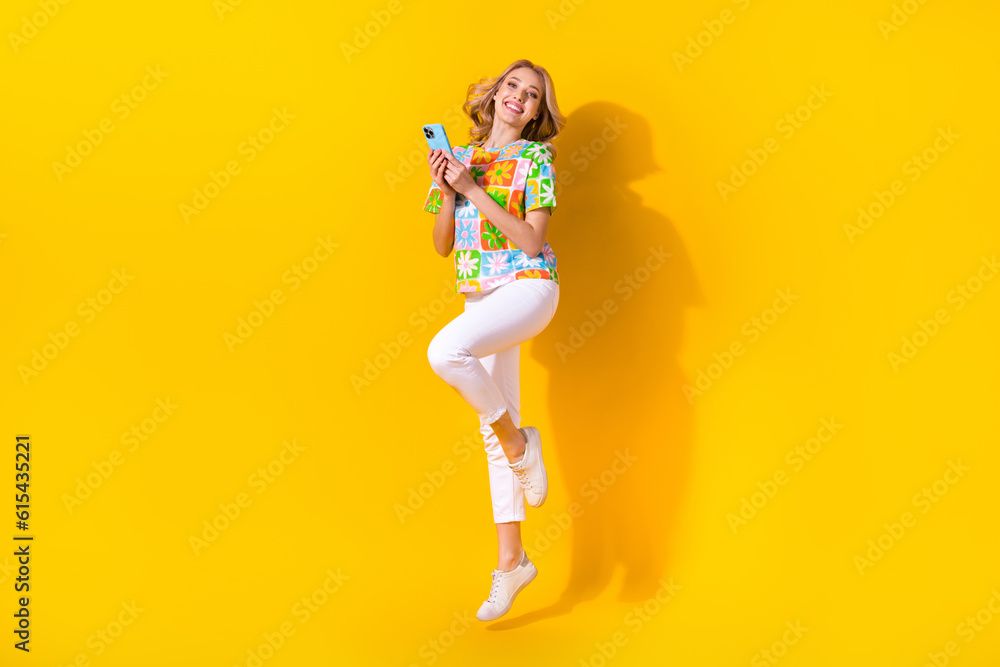 Full length photo of dreamy pretty lady wear flower print t-shirt jumping communicating modern device isolated yellow color background