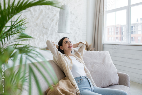 Young self-satisfied female student, business woman sitting on sofa at home and resting. Relaxed and dreamy, she threw her hands behind her head.
