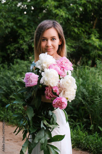 A cute woman is holding bouquet of peonies flower and smiling in the park.