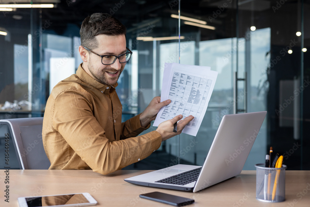 Fototapeta premium A young smilling businessman conducts an online business meeting. He sits in the office at the table in front of the laptop and communicates via video call, shows documents and reports on the screen