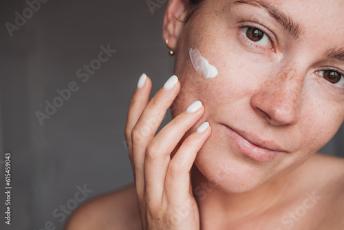Wallpaper Mural Young woman with cream on her cheek touching her face and looking at the camera, beauty routine  Torontodigital.ca