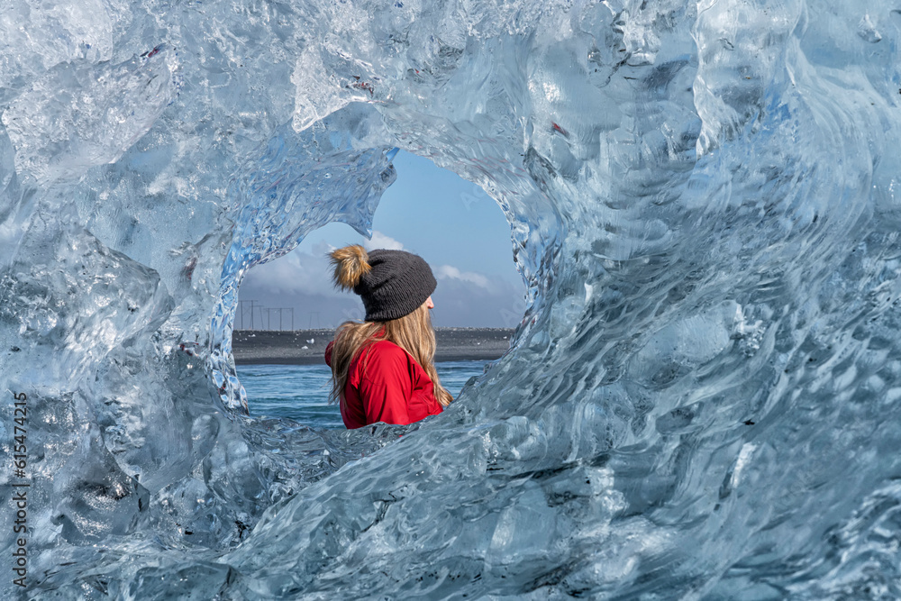 Close-up view through a round, clear ice formation of a woman wearing a woolen hat looking out and admiring the icebergs along the South Coast of Iceland; Jokulsarlon, South Iceland, Iceland Close-up view through a round, clear ice formation of a woman wearing a woolen hat looking out and admiring the icebergs along the South Coast of Iceland; Jokulsarlon, South Iceland, Iceland