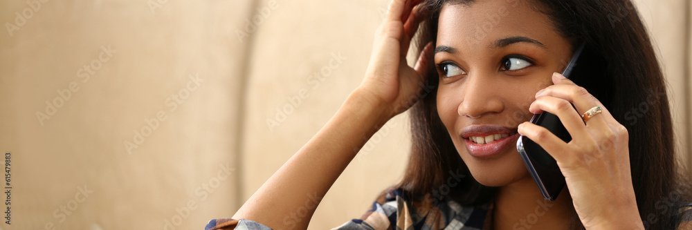Fototapeta premium Smiling pensive young black woman talking on phone in living room. Talking on phone and discussing news