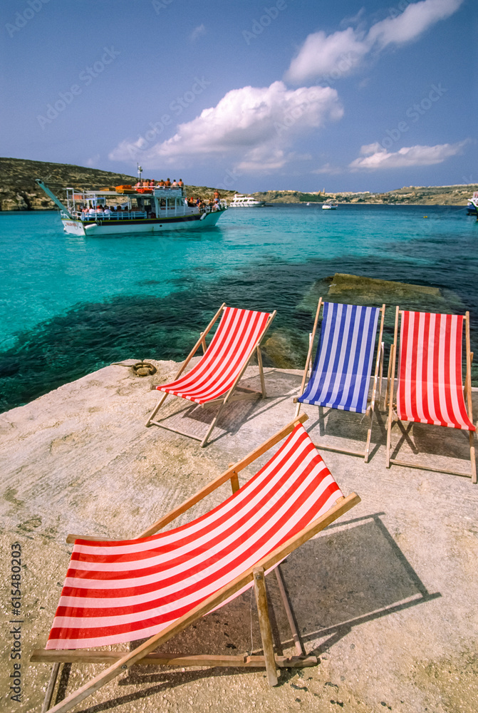 Cruise boat sails by a Mediterranean shoreline with deck chairs; Comino