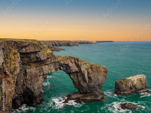 Sunrise over the Green Bridge of Wales. Witness the awe-inspiring beauty of the Green Bridge of Wales at sunrise, with the crashing waves of the sea below and the stunning coast of Pembrokeshire.