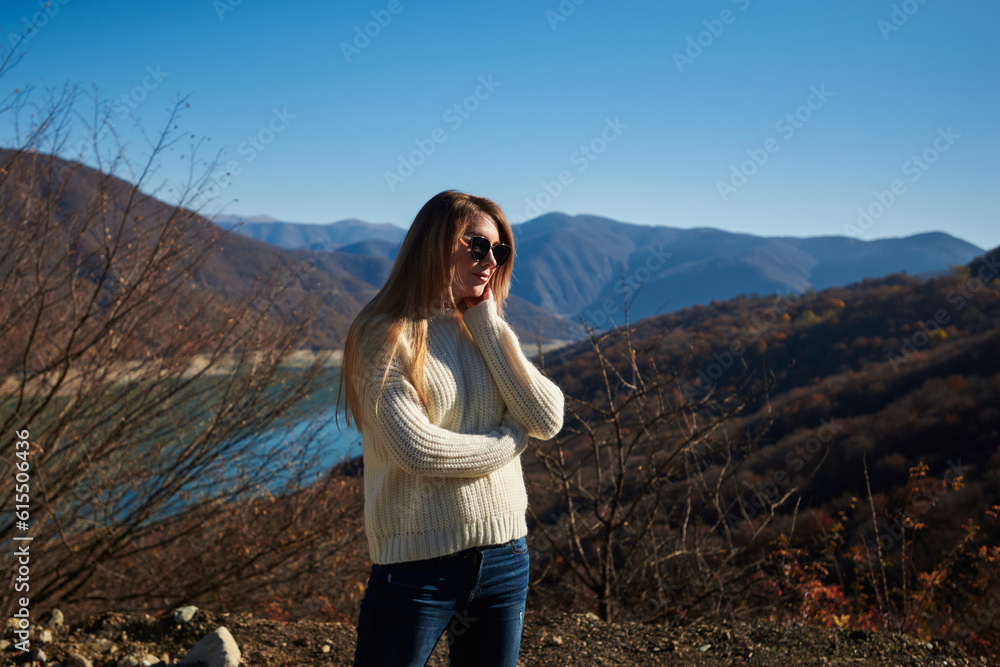 Naklejka premium beautiful young woman with long blond hair in a light sweater and jeans on the shore of an azure lake, autumn trees and mountains