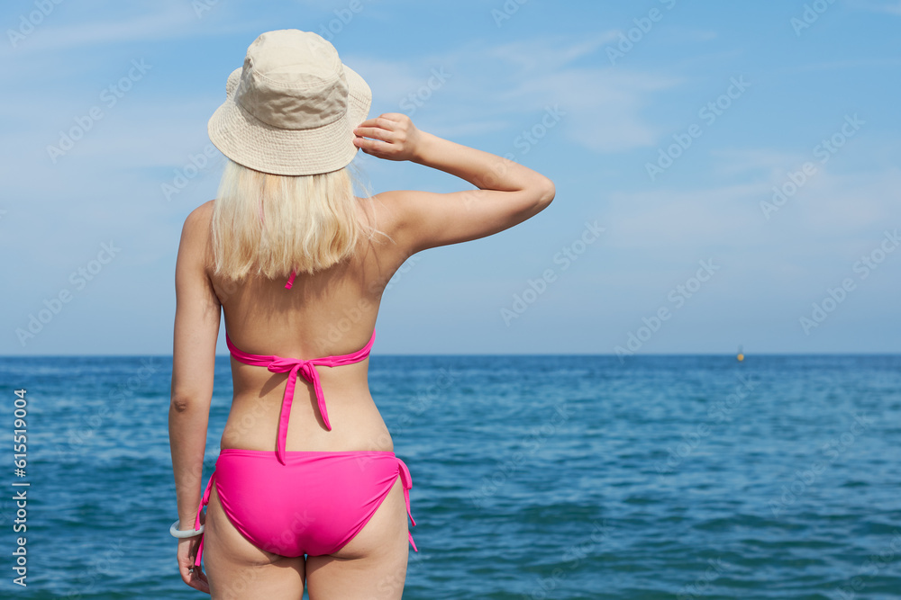 Back view of a blonde girl on the beach with a hat holding it with one hand looking towards the sea on a sunny day.  Girl enjoying the beach in summer. Holidays.