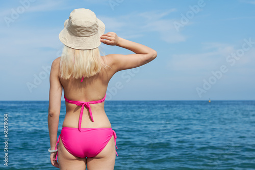 Back view of a blonde girl on the beach with a hat holding it with one hand looking towards the sea on a sunny day. Girl enjoying the beach in summer. Holidays.