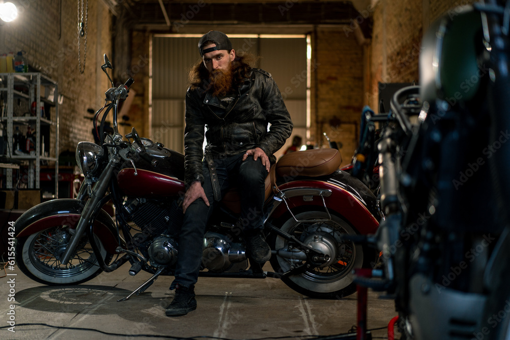 Creative authentic motorcycle workshop Garage brutal serious bearded biker mechanic sitting on motorcycle