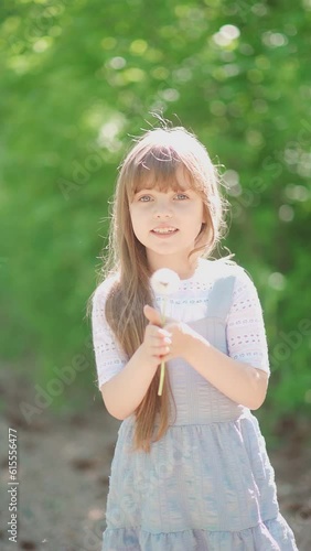 Beautiful cute little girl enjoy blowing dandelion in summer park. Sweet child with spring flower of blowball in midday light. Slow motion. Vertical