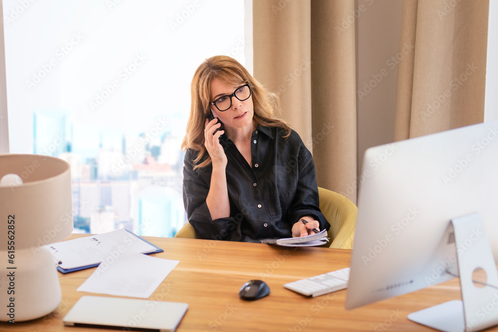 Mid aged businesswoman using computer and mobile phone and working in the office Mid aged businesswoman using computer and mobile phone and working in the office