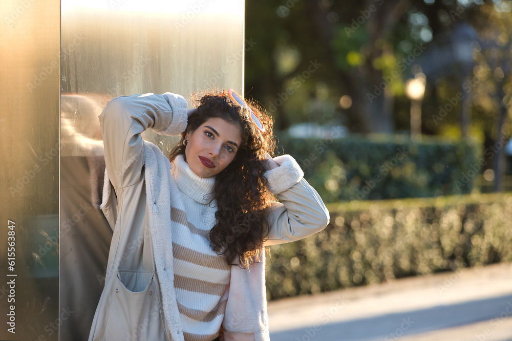 Young woman, beautiful, brunette with curly hair, sweater, coat and sunglasses, leaning on a metal wall, sensual and flirtatious receiving the sun's rays. Concept beauty, fashion, autumn, winter, cold Young woman, beautiful, brunette with curly hair, sweater, coat and sunglasses, leaning on a metal wall, sensual and flirtatious receiving the sun's rays. Concept beauty, fashion, autumn, winter, cold