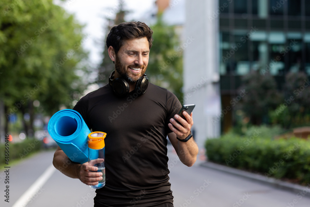 Fototapeta Portrait of mature successful adult athlete, bearded man going to workout, coach holding fitness mat and phone, browsing online app for joint physical training