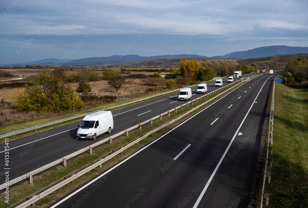 A large number of minivans move along the highway carrying goods for all people in the world. White modern delivery small shipment cargo courier van moving fast on motorway road to city urban suburb. 