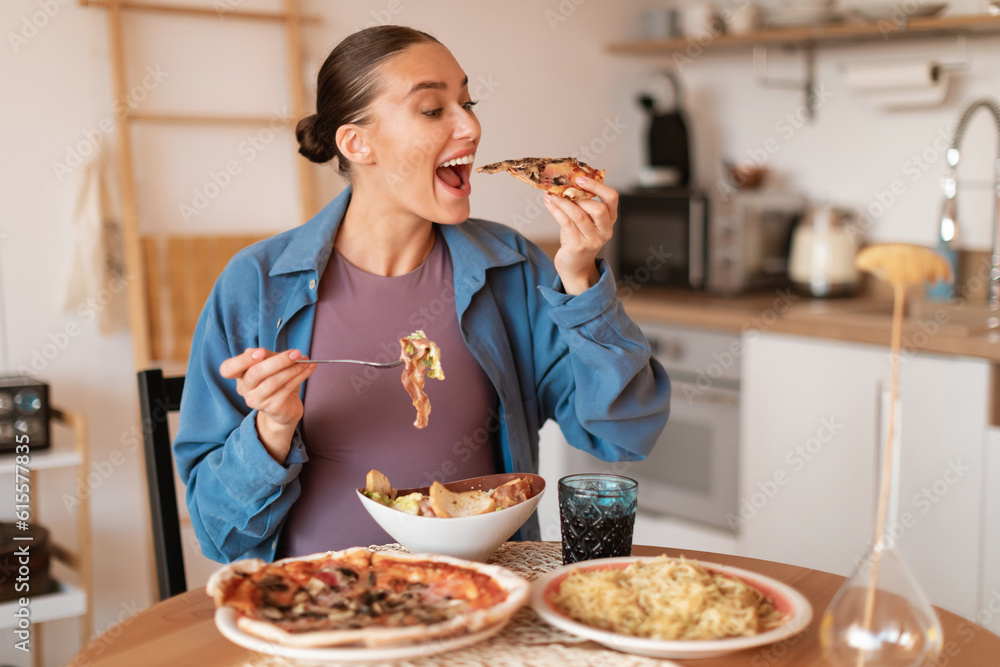 Expectant delights. Hungry pregnant woman eating pizza and salad, having desire for junk food, sitting in kitchen Expectant delights. Hungry pregnant woman eating pizza and salad, having desire for junk food, sitting in kitchen