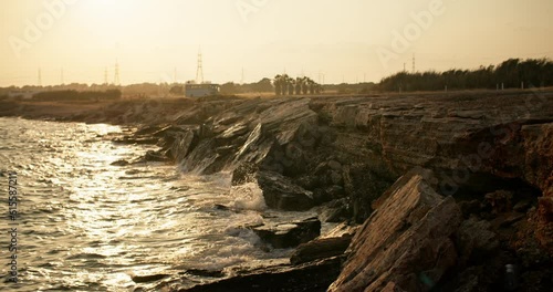 A Captivating Spectacle: Waves of the Sea Embrace the Sun's Last Glow, Cascading Against the Ancient Coastal Rocks at Sunset. High quality 4k footage