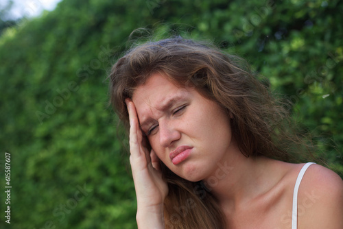Portrait of frustrated sad upset young woman on natural green background suffering from headache, migraine, feel strong pain