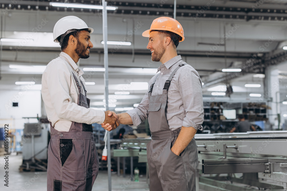 Two international male workers in overalls shake hands against the background of the production of PVC windows and doors