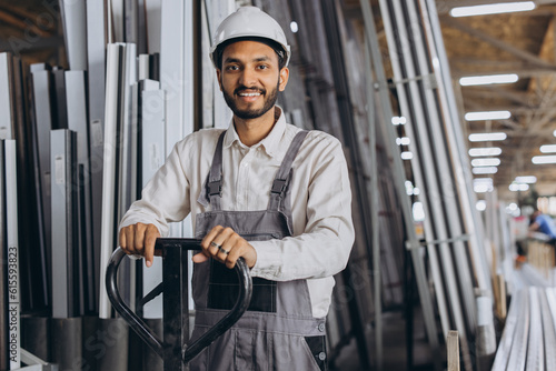 Portrait of a happy Hindu worker in a white hard hat and overalls holding a hydraulic truck against a background of a factory and aluminum frames.