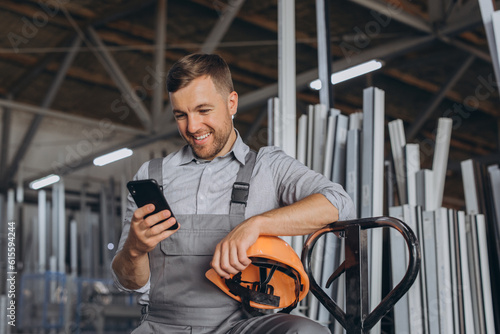Portrait of a happy worker in a orange helmet and overalls holding a hydraulic truck and talking on the phone against a background of a factory and aluminum frames. © anatoliycherkas