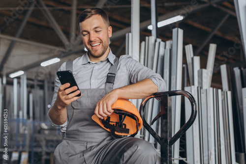 Portrait of a happy worker in a orange helmet and overalls holding a hydraulic truck and talking on the phone against a background of a factory and aluminum frames.