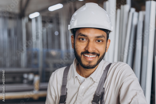 Portrait of a happy Hindu worker in a white hard hat and overalls holding a hydraulic truck against a background of a factory and aluminum frames. © anatoliycherkas