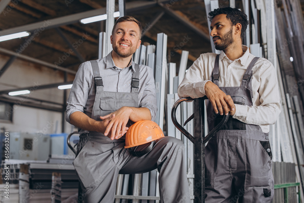 Portrait of two international workers wearing hardhats taking break from work and resting speaking to each other on a factory background with copy space Portrait of two international workers wearing hardhats taking break from work and resting speaking to each other on a factory background with copy space