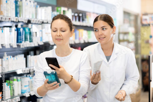 Woman pharmacist in medical uniform helps woman shopper in casual clothes find cosmetic product from photo on internet