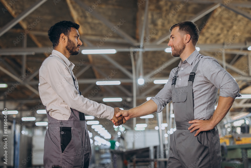 Two happy international workers in overalls shake hands against a factory background Two happy international workers in overalls shake hands against a factory background