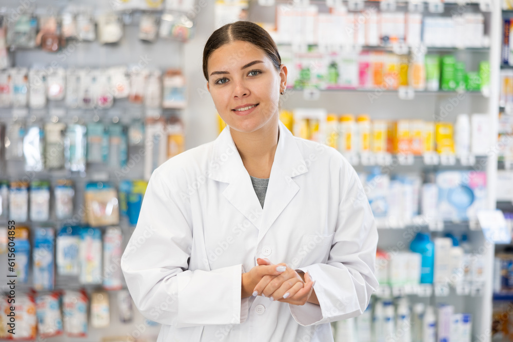 Young female pharmacist in uniform posing against backdrop of shelves with products in pharmacy