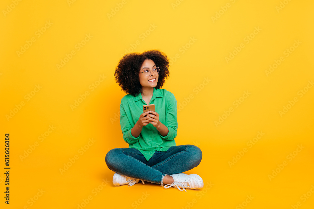 Full length photo of happy positive trendy african american or brazilian curly woman, sits on isolated yellow background, uses smartphone, chats on social networks, smiles, looks away, dreaming Full length photo of happy positive trendy african american or brazilian curly woman, sits on isolated yellow background, uses smartphone, chats on social networks, smiles, looks away, dreaming